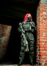 Airsoft red-hair woman in uniform with machine gun standing on ruins. Vertical photo
