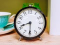 An alarm clock with a black case and black numbers and arrows on a white dial stands on a wooden table