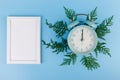 Alarm clock, blank white photo frame, cypress branches on blue background. Christmas concept Royalty Free Stock Photo