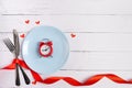 Alarm clock on blue plate with red hearts, table knife and fork, wooden background with copy space