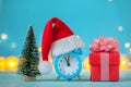 Alarm clock with Christmas Santa hat and christmas tree and present box on blue background. Time for Christmas. Royalty Free Stock Photo