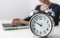 An alarm clock on a desk is the focus with a person typing in the blurred background, Time management, work environment, office
