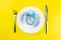 Alarm clock with fork and knife on the white plate on yellow table. Time to eat, Breakfast, Lunch Time and Dinner concept Royalty Free Stock Photo