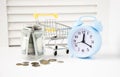 Alarm clock and mini shopping cart full of coins on wooden table, natural green blurred background. Royalty Free Stock Photo