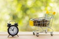 Alarm clock and mini shopping cart full of coins on wooden table, natural green blurred background. Business, finance, time,