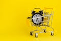 Alarm clock in mini shopping cart on yellow background. Buying, time, sale concept.