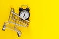 Alarm clock in mini shopping cart on yellow background. Buying, time, sale concept. Flat lay, top view