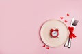 Alarm clock on pink plate with red hearts, table knife and fork, background with copy space