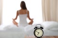 Alarm clock standing on bedside table has already rung early morning to wake up woman in bed sitting in background