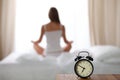 Alarm clock standing on bedside table has already rung early morning to wake up. Woman do yoga in bed in background