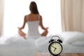 Alarm clock standing on bedside table has already rung early morning to wake up. Woman do yoga in bed in background