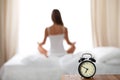 Alarm clock standing on bedside table has already rung early morning to wake up. Woman do yoga in bed in background