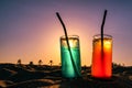 Alcoholic coctail drinks with a straw and ice in the sand with palm trees and colorful sky at background