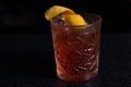 Alcoholic drink of red color with ice in beautiful engraved glass decorated with lemon zest. Standing on bar stand surface. Side