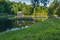 An alcove on the water, a bridge leading to it, green grass and a lake in the foreground