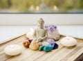 All seven chakra colors crystals stones around sitting Buddha figurine on natural wooden tray.
