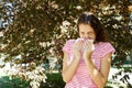 Allergy concept. Sneezing young girl with nose wiper among blooming trees in park Royalty Free Stock Photo