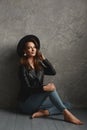 Alluring model girl with slim body in a leather jacket, stylish hat and jeans posing on the wooden floor indoors