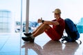 Alone backpacker traveller sitting on the airport terminal floor, looking at hall window and waiting for boarding at aircraft