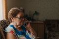 Alone elderly woman talking on cell phone near the window in her living room. Communication concept