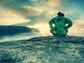 Alone girl sits with her back on cliff against huge misty mountain