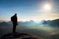 Alone photographer with heavy backpack and tripod in hand on rocky cliff and watching down to deep misty valley bellow