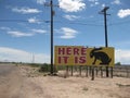 Along a desert road, a yellow billboard sign reads Here It Is with a rabbit's silhouette