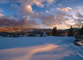 Alpine village outskirts in last evening sunset sun light. Winter snowy hills and fir trees. Picturesque clouds and Moon in dusk