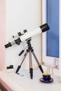 An amateur astronomical telescope stands on a tripod and is pointed out the window