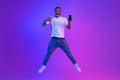 Amazed African Man Jumping Over Purple Neon Background And Pointing At Smartphone