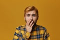 Amazed and astonished male wears checkered yellow shirt with beard and dyed red hair bieng shocked.