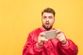 Amazed bearded man with a smartphone in his hands, looks at the camera with an expressive look, wears a red jacket, stands on a Royalty Free Stock Photo