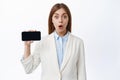 Amazed ceo office woman in business suit shows mobile screen, holding phone horizontally and demontrate empy display