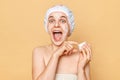 Amazed excited woman in bath cap and wrapped in towel posing after taking shower applying face cream doing cosmetology procedures Royalty Free Stock Photo