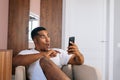 Amazed African-American man sitting at comfortable armchair and using mobile phone. Royalty Free Stock Photo