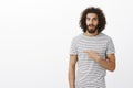 Amazed handsome musculine guy with beard and curly hair in trendy striped t-shirt, pointing right and teeling advice to