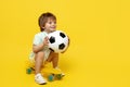 Amazed little boy play with soccer ball while sitting on pennyboard or skateboard over yellow background.