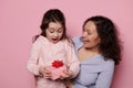 Happy little girl expressing surprise while getting birthday present from her loving mother, isolated on pink background