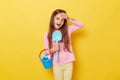 Amazed little girl wearing casual clothing holding sandbox toys and bucket isolated over yellow background looking at something Royalty Free Stock Photo