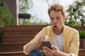 Amazed man with open mouth using mobile phone shopping online sitting on the street. Emotional freelancer reading good news, recei