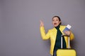 Amazed woman wearing green pull and yellow jacket, standing near suitcase and pointing at copy space on gray background Royalty Free Stock Photo