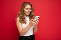 Amazed shocked beautiful young blonde woman wearing white t-shirt isolated over red background using smartphone and