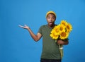 Amazed, shocked young African American man with armful of sunflowers pointing with hand up wearing green t-shirt and yellow hat