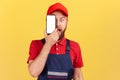 Amazed worker man in blue uniform standing and covering eye with cell phone with empty display.