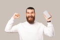 Amazed young bearded man in white hoodie celebrating success and holding smartphone with blank screen Royalty Free Stock Photo