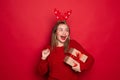 Amazed young Santa girl in fun decorative deer horns on head pointing index finger on red boxes with gifts presents on red Royalty Free Stock Photo