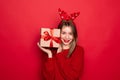 Amazed young Santa girl in fun decorative deer horns on head pointing index finger on red boxes with gifts presents on red Royalty Free Stock Photo
