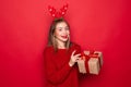 Amazed young Santa girl in fun decorative deer horns on head pointing index finger on red boxes with gifts presents on red Royalty Free Stock Photo