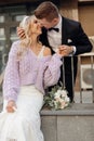 Amazing couple of young bride with long wavy fair hair in bridal dress, lilac cardigan hugging and kissing bridegroom.