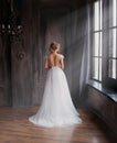 Amazing lady in long white adorable expensive light dress with train and open back stands with back to camera, girl with